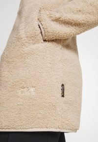 Beżowa polarowa kurtka o miękkiej, teksturowanej powierzchni. Posiada naszywkę z logo oraz metkę na bocznym końcu, ma luźny krój i długie rękawy.