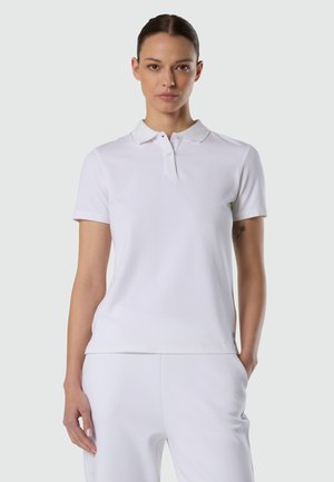 Donna con capelli scuri che indossa una polo bianca a maniche corte e pantaloni bianchi, in piedi con una mano in tasca davanti a uno sfondo chiaro e semplice.