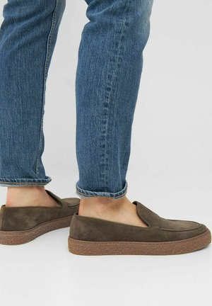 Zapatos slip-on de ante marrón con una suela de goma plana, diseñados con una punta redondeada y sin cierres visibles, combinados con unos jeans de mezclilla azul con dobladillo.