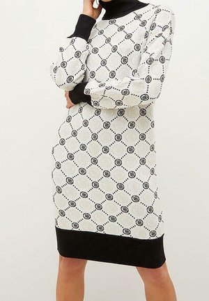 Kobieta ubrana w białą dzianinową sukienkę do kolan z czarnym okrągłym wzorem, czarnymi mankietami i czarnym dołem, stojąca z jedną ręką przy twarzy.