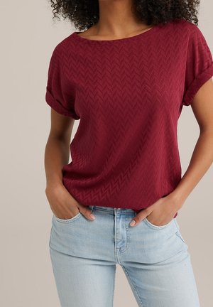 Bordeauxrotes Kurzarmoberteil mit strukturiertem Zickzackmuster, weitem Ausschnitt und lockerem Schnitt, kombiniert mit hellblauen Skinny-Jeans.