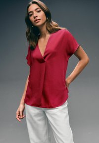 Fuchsia v-neck top with short cap sleeves, made of smooth fabric. Paired with white pants, features a relaxed fit and subtle stitching.