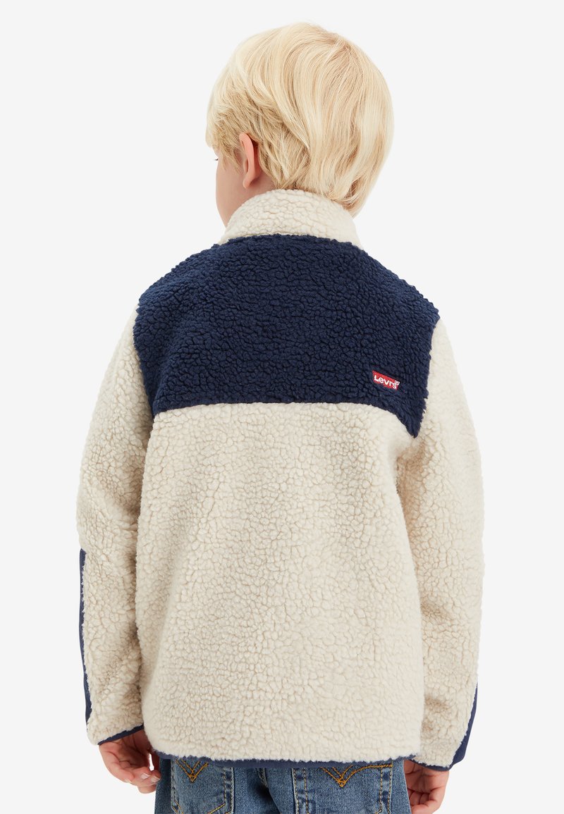Chaqueta de forro polar suave en color crema con una sección superior azul marino. Presenta un cuello alto y un pequeño logo rojo en la espalda. Tejido texturizado.