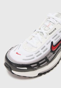 Bela športna obutev z črnimi in sivo akcenti, z mrežasto zgornjim delom, vezalkami in rdečim Nike logotipom. Max Air oblazinjenje je vidno v podplatu.