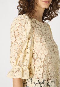Krajková blůza v jemné žluté barvě s květinovými vzory, nabírané rukávy a volánkové manžety. Poloprůhledná textura zvýrazňuje designové detaily.