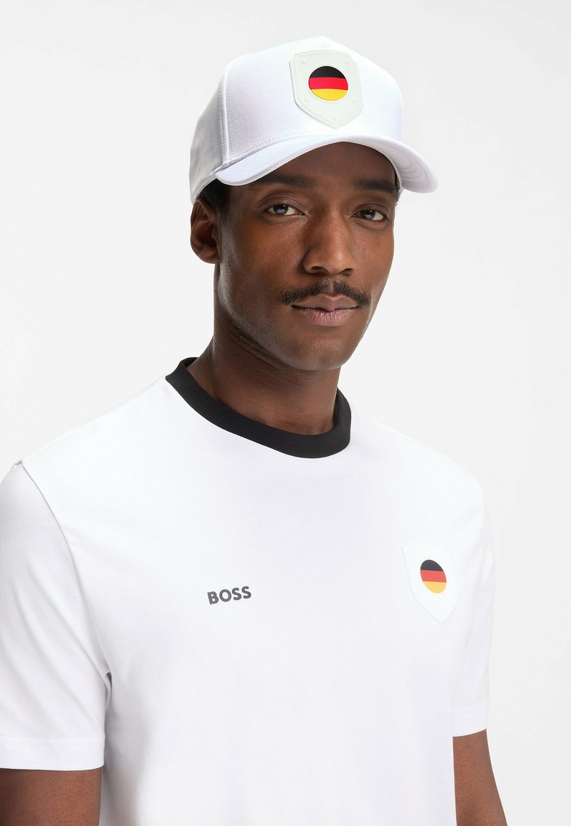 Homme portant une casquette blanche et une chemise, toutes deux ornées de patchs du drapeau allemand et du logo "BOSS" sur la poitrine de la chemise.