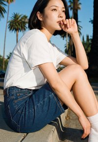 Tânără în tricou alb și fustă de denim, așezată pe bordură, sprijinindu-și bărbia în mână, cu palmieri și cer senin în fundal.