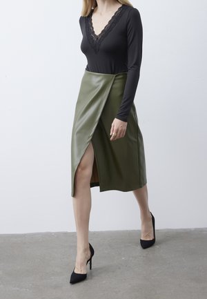 Haut noir à manches longues avec décolleté en V en dentelle, associé à une jupe portefeuille en simili cuir olive taille haute avec une fente sur le côté. Escarpins noirs.