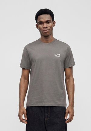 Jeune homme aux cheveux courts et bouclés portant un t-shirt gris uni EA7 Emporio Armani et un pantalon sombre, debout sur un fond blanc.