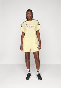 Žltá športová súprava pozostávajúca z trička s krátkym rukávom a kraťasov. Čierne pruhy Adidas na rukávoch a logo v ružovej farbe. Čierne topánky s bielymi pruhovanými ponožkami.