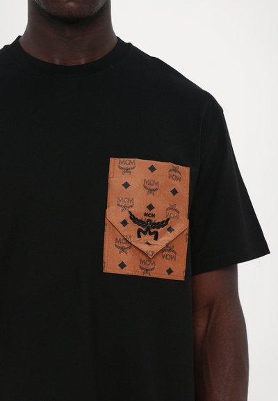 T-shirt noir avec une poche marron ornée d'un motif répétitif et d'un logo. La poche présente une finition texturée et des bords nets.