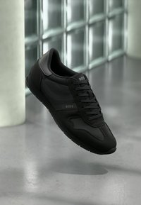 Schwarzer Sportschuh mit glattem Lederobermaterial, strukturierten Akzenten und Schnürdesign. Verfügt über dezente Logo-Details an der Seite.