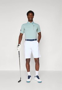 Golfantrekk med lys blå polotrøye med korte ermer, en glidelåslomme, hvite shorts og hvite sko med blå detaljer.