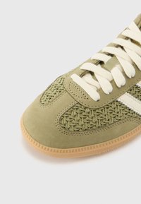 Grøn ruskinds sneaker med vævet tekstur, cremefarvede snørebånd og en beige gummisål med et struktureret mønster på bunden.