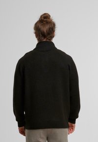 Zwarte, chunky gebreide trui met een hoge, geribbelde kraag en afgezakte schouders. Heeft lange mouwen en een relaxte pasvorm, gemaakt van zacht materiaal.