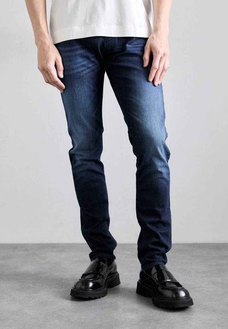Dunkelblaue Jeans mit schmaler Passform und einem verwaschenen Finish. Kombiniert mit glänzenden schwarzen Schuhen und einem Oberteil in neutralen Farben. Einfache Designdetails.