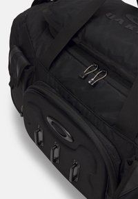 Mustapukuinen duffel-laukku, jossa on teksturoitu kangas, kaksinkertaiset vetoketjusangat, kolme mustaa metallikoukkua ja korotettu logo etupaneelissa.