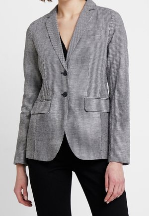 Femme portant un blazer cintré noir et blanc à petits carreaux, avec deux boutons et des poches avant à rabat, par-dessus un pantalon noir.