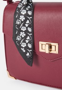 Borsa in pelle bordeaux con chiusura dorata, dotata di una sciarpa nera a fantasia floreale annodata al manico. Texture liscia, design strutturato.