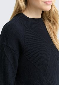 Ciemnoniebieski sweter dzianinowy z teksturowanym wzorem, z prążkowanym dekoltem i wyraźnymi ukośnymi liniami. Widoczny miękki, przytulny materiał.