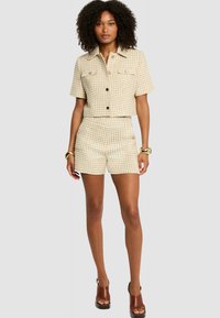 Chemise à manches courtes, crop, en tartan avec deux poches poitrine, associée à un short taille haute. Le tissu est beige avec des accents marron et une finition texturée.