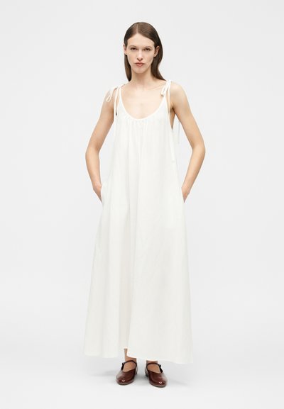 Γυναίκα με ίσια καστανά μαλλιά, που φοράει μακρύ λευκό φόρεμα χωρίς μανίκια με ιμάντες δέσιμο και καφέ παπούτσια με κλειστή μύτη, όρθια με τα χέρια στις τσέπες.