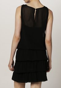 Musta ilmanhiha dressi kerroksellisella rypytyksellä, läpinäkyvällä takapaneelilla ja pienellä nappikiinnityksellä kaulassa. Sujuva pinta.