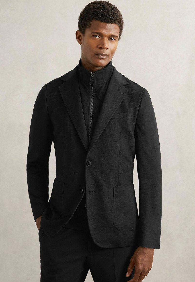 Sort skræddersyet jakke med slim fit, der har en notch revers, to forlommer og et lynlås indre lag. Glat tekstur, ingen synlige mønstre.