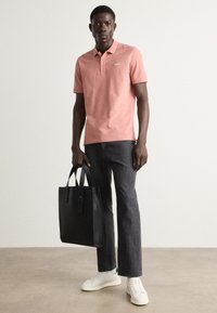 Chandail polo rose à manches courtes avec un logo discret, associé à un jean foncé et des baskets blanches ; portant un sac fourre-tout en cuir noir.