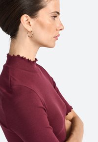 Haut bordeaux à côtes avec un col haut en volants et des manches courtes. Le modèle porte des boucles d'oreilles en or. Tissu texturé.