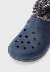 Marineblå slip-on klogs med perforerede huller og blødt gråt fleecefôr, designet til varme og åndbarhed.
