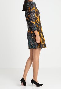 Robe à manches courtes avec un motif cachemire orange, noir et bleu sarcelle, dotée d'un corsage cintré et d'une jupe évasée. Associée à des escarpins noirs à talons aiguilles.