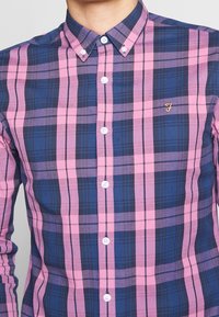 Blå och rosa rutig skjorta med knappar som har spridd krage, vita knappar och en liten guldsymbol på vänstra bröstet. Mjuk tygtextur.