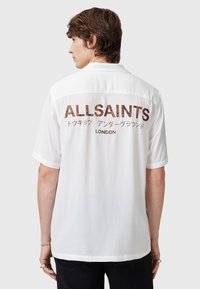 Witte korte mouwen overhemd met "ALLSAINTS" in bruine patroonletters op de achterkant, met een klassieke kraag en een gladde textuur.