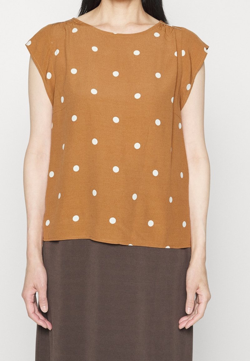Blusa color ruggine con pois bianchi, maniche corte, scollatura rotonda e vestibilità leggermente ampia, abbinata a un pantalone marrone scuro.