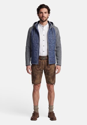 Mann trägt blaue gesteppte Jacke mit grauen Ärmeln, weißes Hemd, traditionelle braune Lederhose, beige Socken und braune Schuhe, steht nach vorne gewandt.