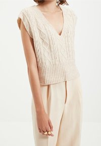 Gilet di maglia beige con scollatura a V, senza maniche e motivo a trecce. Presenta un orlo a costine ed è abbinato a pantaloni di colore chiaro.