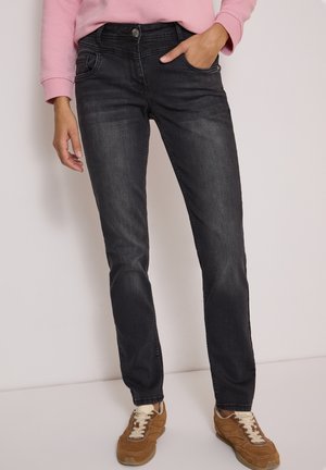 Personne portant un jean slim noir, des baskets marron à lacets et un haut rose à manches longues, une main dans la poche.