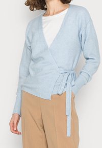 Kobieta w jasnoniebieskim swetrze zapinanym w talii, noszącym się na białej bluzce, w połączeniu z beżowymi spodniami, stoi z jedną ręką w kieszeni.