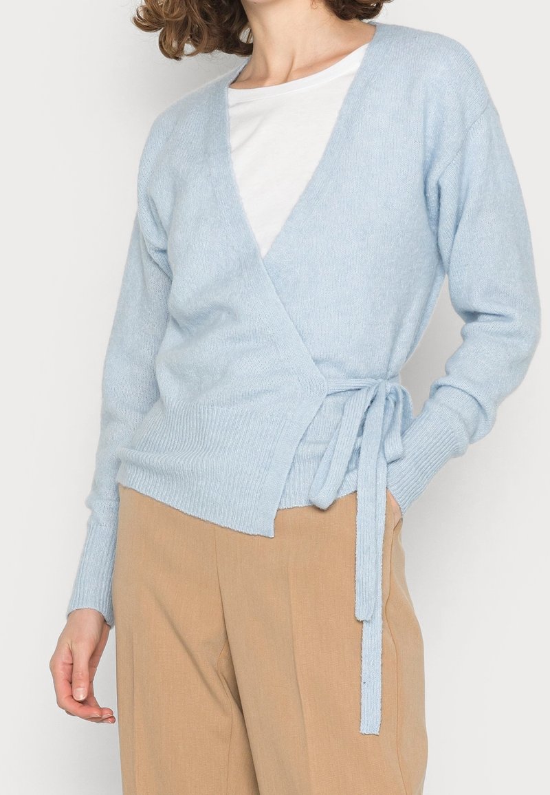 Kobieta w jasnoniebieskim swetrze zapinanym w talii, noszącym się na białej bluzce, w połączeniu z beżowymi spodniami, stoi z jedną ręką w kieszeni.