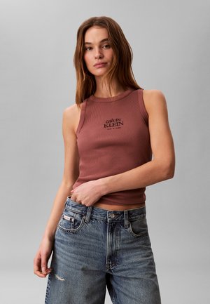 Giovane donna che indossa un top senza maniche color bordeaux di Calvin Klein e jeans larghi blu, davanti a uno sfondo grigio uniforme.