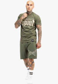 Zelené bavlnené tričko s bielym vintage štýlovým logom "Tapout" a zodpovedajúce šortky s rovnakým logom. Na sebe má biele tenisky a pruhované ponožky.