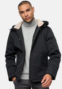 Chaqueta de invierno negra con capucha forrada de piel sintética, cierres de cremallera y botón a presión, bolsillos frontales dobles y un tejido texturizado. Llevada sobre un suéter de cuello alto gris.