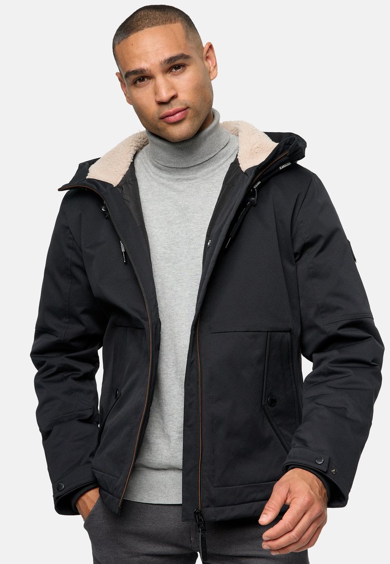 Chaqueta de invierno negra con capucha forrada de piel sintética, cierres de cremallera y botón a presión, bolsillos frontales dobles y un tejido texturizado. Llevada sobre un suéter de cuello alto gris.