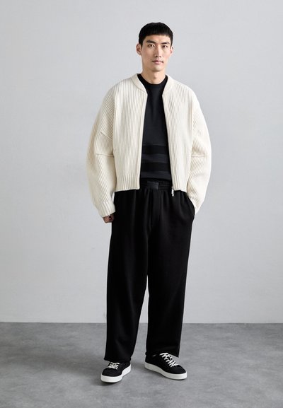 Weißer gerippter Cardigan über einem schwarzen Shirt, kombiniert mit lockeren schwarzen Hosen und schwarzen Sneakers. Minimalistisches Design mit strukturierter Silhouette.