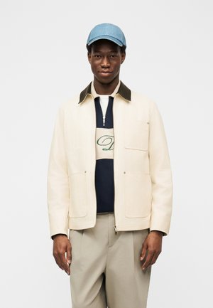 Mlad moški, oblečen v svetlo kremno jakno preko mornarsko modrega in belega puloverja z zadrgo, bež hlače in svetlo modro baseball kapo, stoji ob belem ozadju.