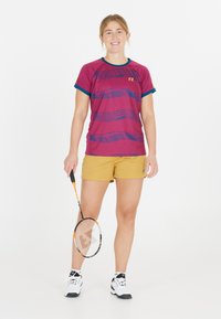 Mörkröd idrottströja med blå detaljer och vågmönster, i kombination med gula shorts. Tillbehör: badmintonracket, vita skor med svarta sulor.