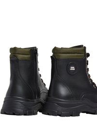 Botas de tobillo de cuero negro con cuello de textil verde, suela de goma, cremallera lateral y ojales de metal. Parche con el logo en la parte trasera.
