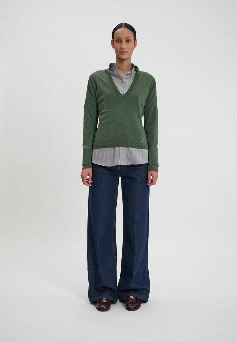 Un pull vert en V porté par-dessus une chemise grise à rayures, associé à un jean foncé à pattes larges. Des chaussures marron brillantes complètent le look.