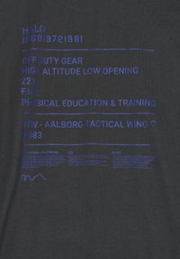 Melna T-kreklā, kurā ir zils drukāts teksts, kas norāda "Halo," "Off Duty Gear" un citas ar militāro tēmu saistītas frāzes ar diagrammu akcentiem.